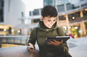 A woman smiling on two digital devices, a smart phone and tablet.