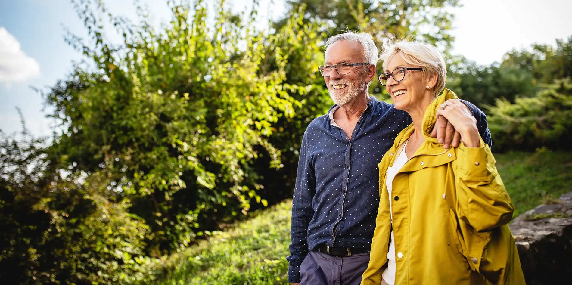 An couple, smiling and embracing, walks outdoors. The man wears a blue shirt, and the woman wears a yellow jacket. They're surrounded by greenery.